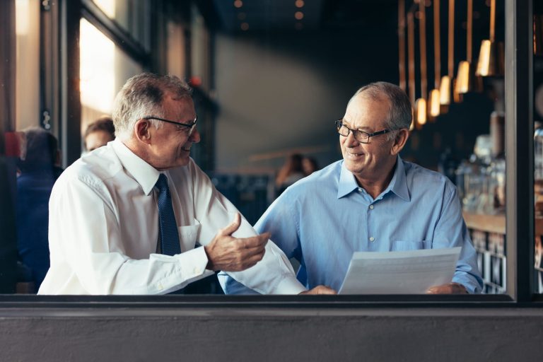 Senior business man with male colleague working together in modern restaurant. View from through window glass of two mature business partners working together at a cafe.