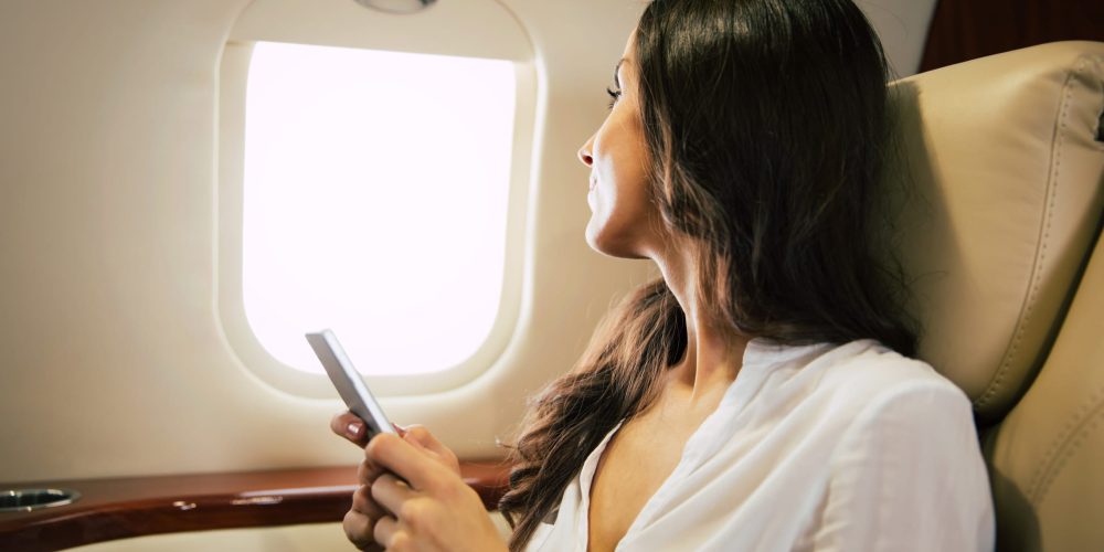 Gorgeous businesswoman in formal attire, who is looking through the window and holding a smartphone, while sitting in her window seat in a business class plane.
