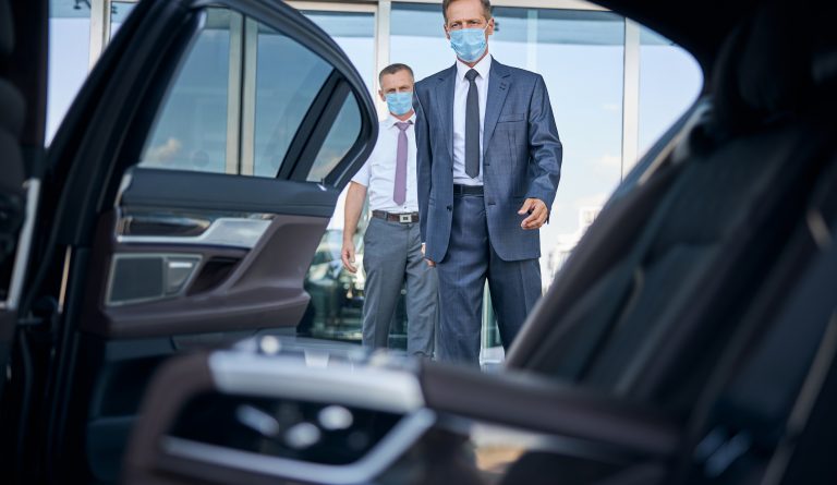 Elegant mature man in sterile mask is getting into automobile after arrival while chauffeur is holding suitcase